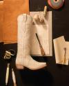 A white cowboy boot lies on a dark surface surrounded by leather, thread spools, handwritten letters, a leatherworking tool, and crafting materials.
