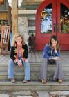 Two women sit on the steps of a wooden porch with red double doors behind them, flanked by decorative statues and rocking chairs.