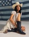Woman in a white dress and cowboy hat sits in front of a large American flag backdrop, wearing black cowboy boots with star designs.