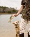 Person holding a small fish by the shore of a lake, with a dog standing in the shallow water nearby.