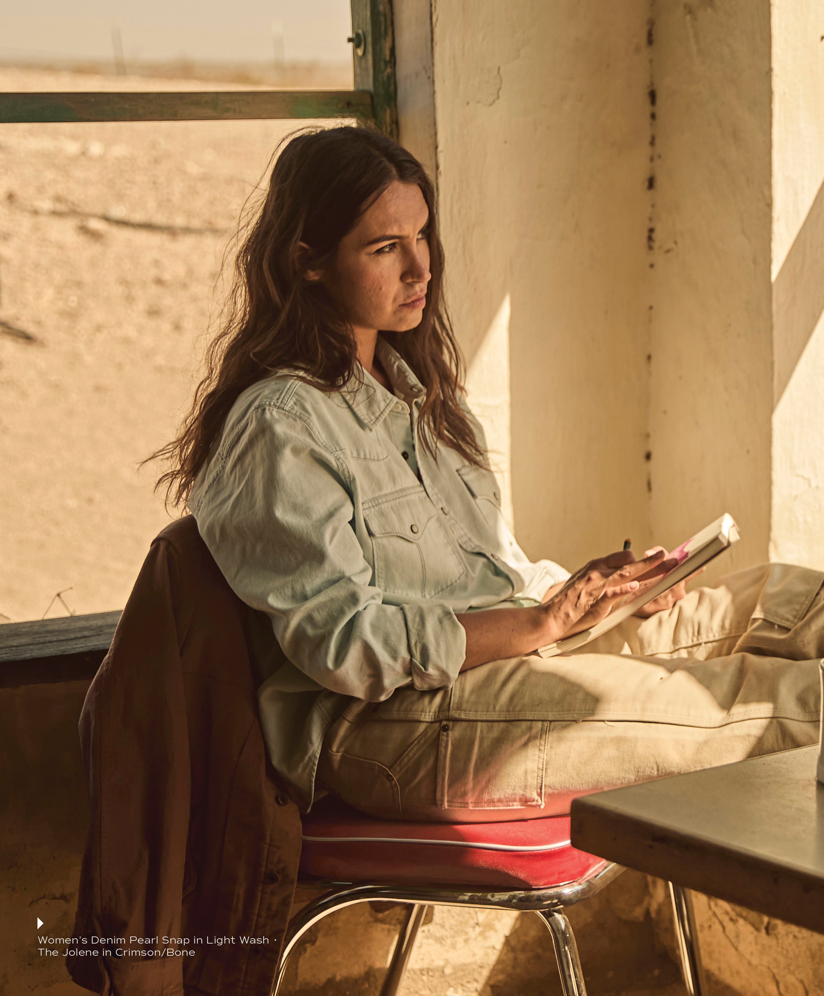 A woman sitting indoors by a sunny window in a light wash Women's Denim Pearl Snap shirt and tan cargo pants, holding a notebook and pen.