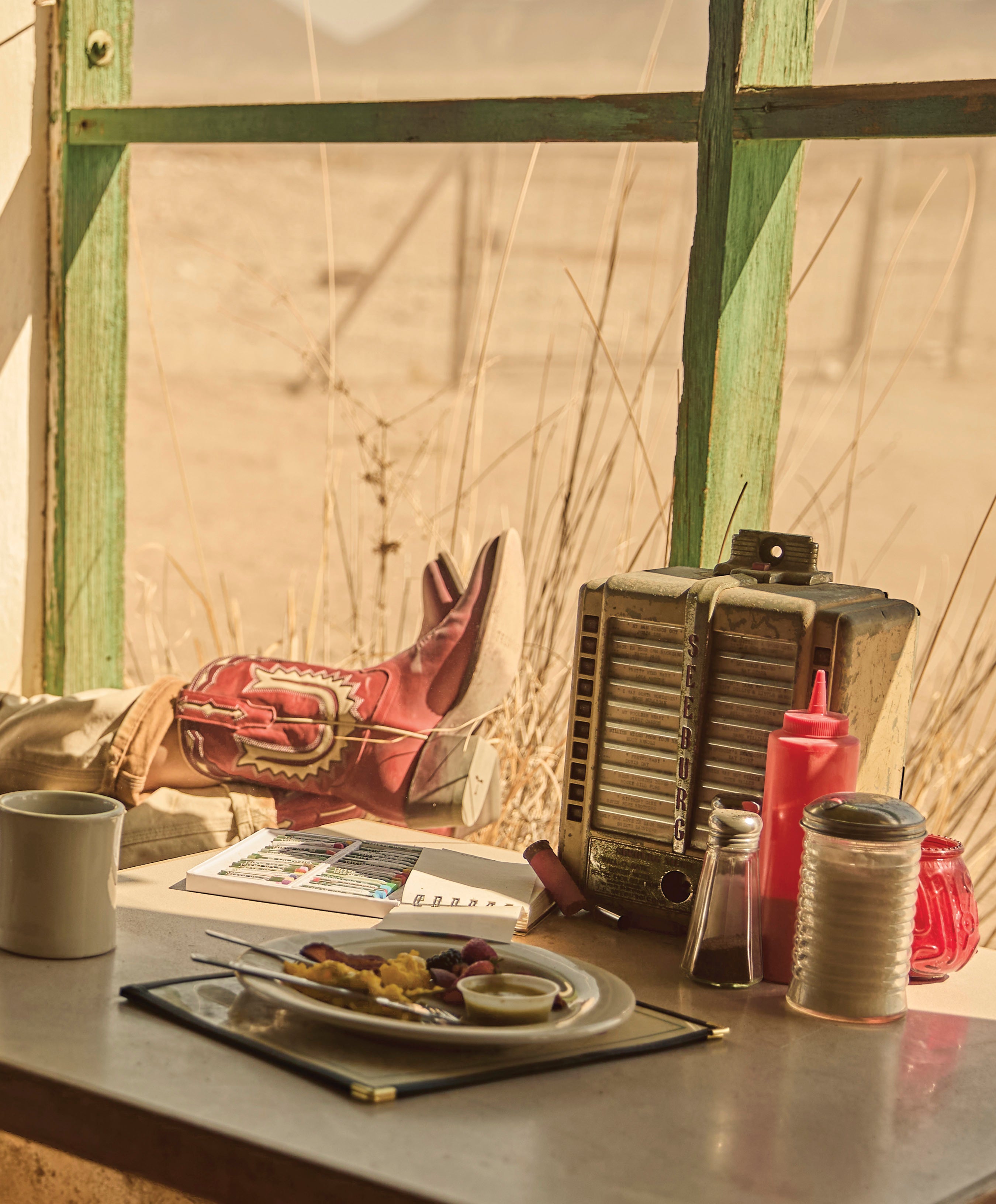 A close-up of a table inside a dusty diner with a plate of food, condiments, and a vintage radio. A person's leg is propped on the windowsill, showing a distinctive red cowboy boot with white embroidery.