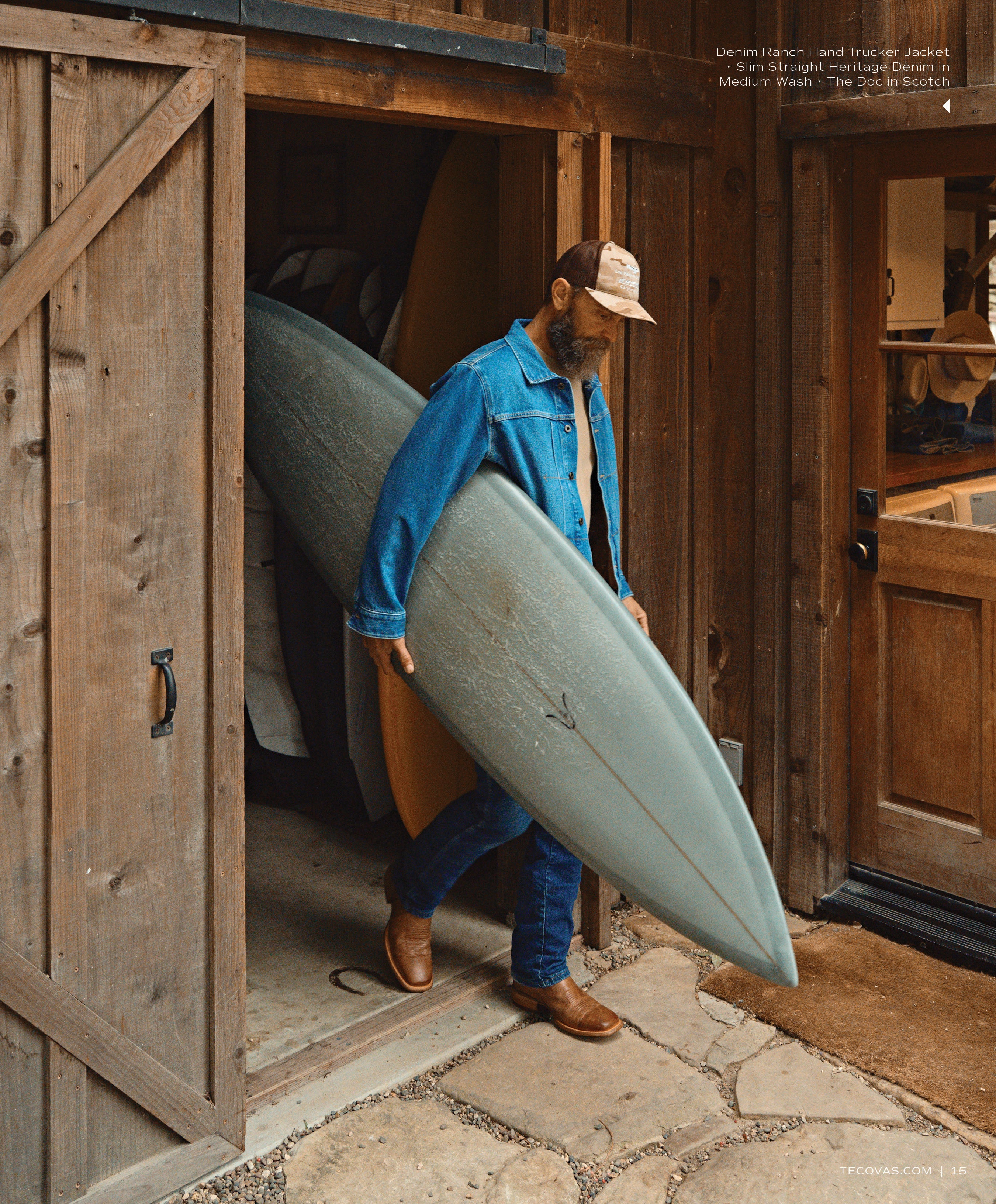 A man with a beard, wearing a denim jacket and a baseball cap, walking out of a rustic wooden barn while carrying a large gray surfboard, wearing brown boots.