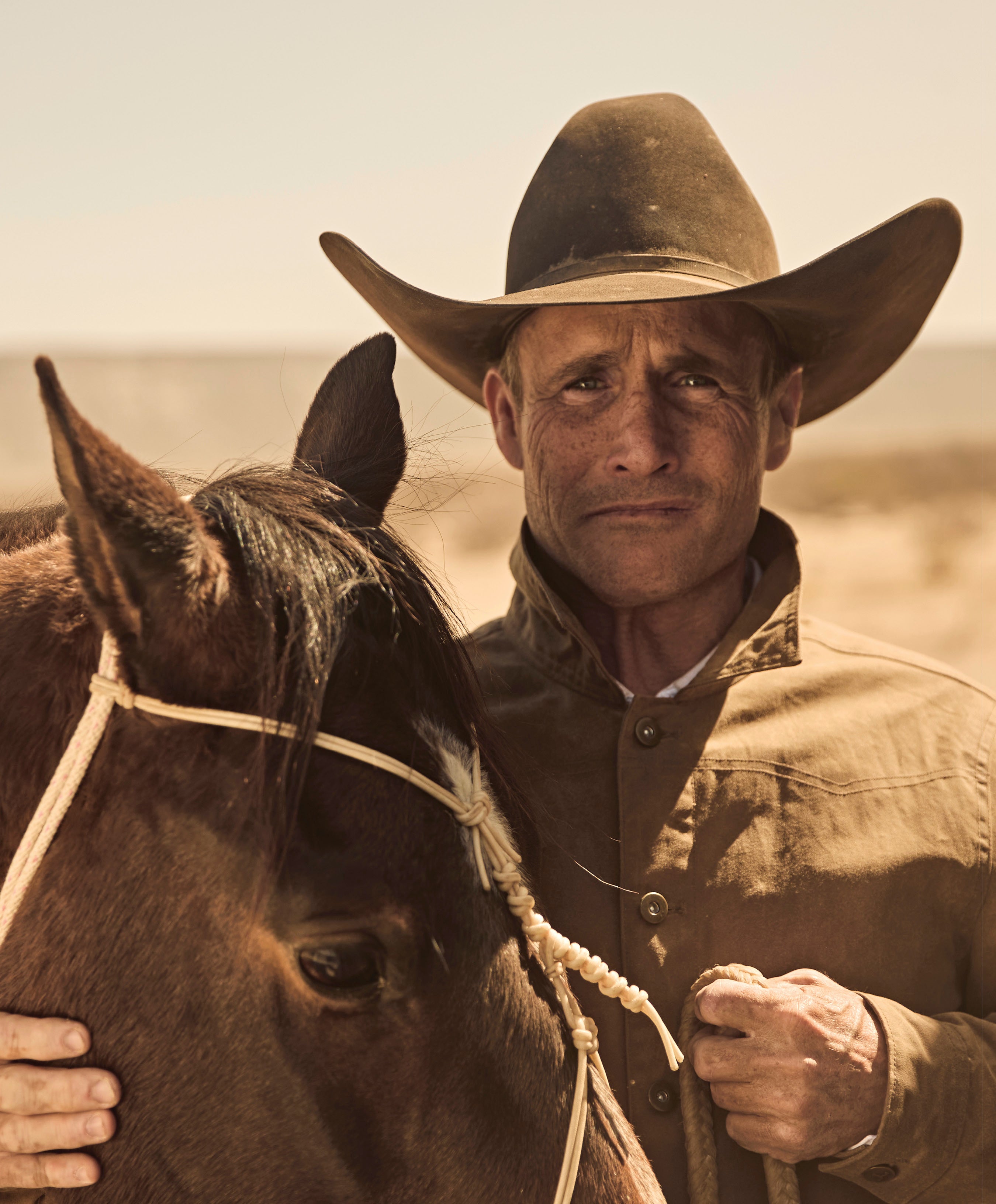 A close-up portrait of a rugged man in a brown cowboy hat and jacket, standing next to a horse in a sunny, dry outdoor setting.