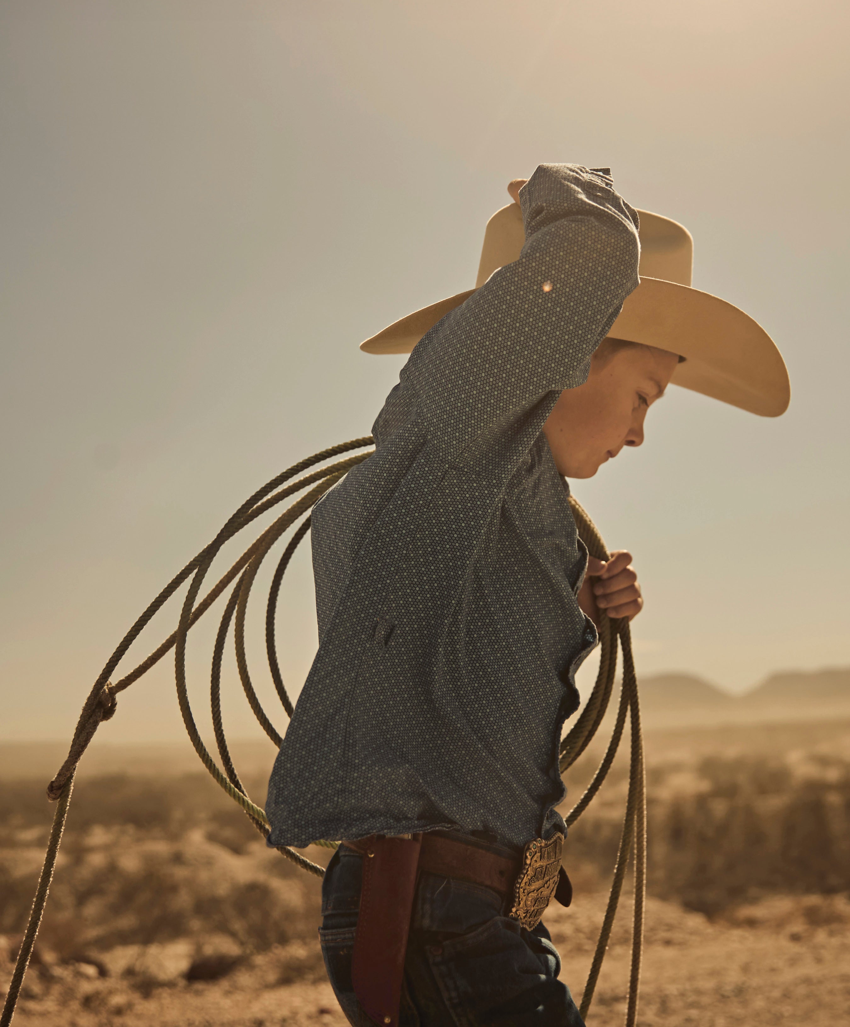 A young cowboy in a light-colored hat, denim shirt, and jeans holding a coiled rope (lariat) over his shoulder, silhouetted against a bright, dry, sunny, desert landscape.