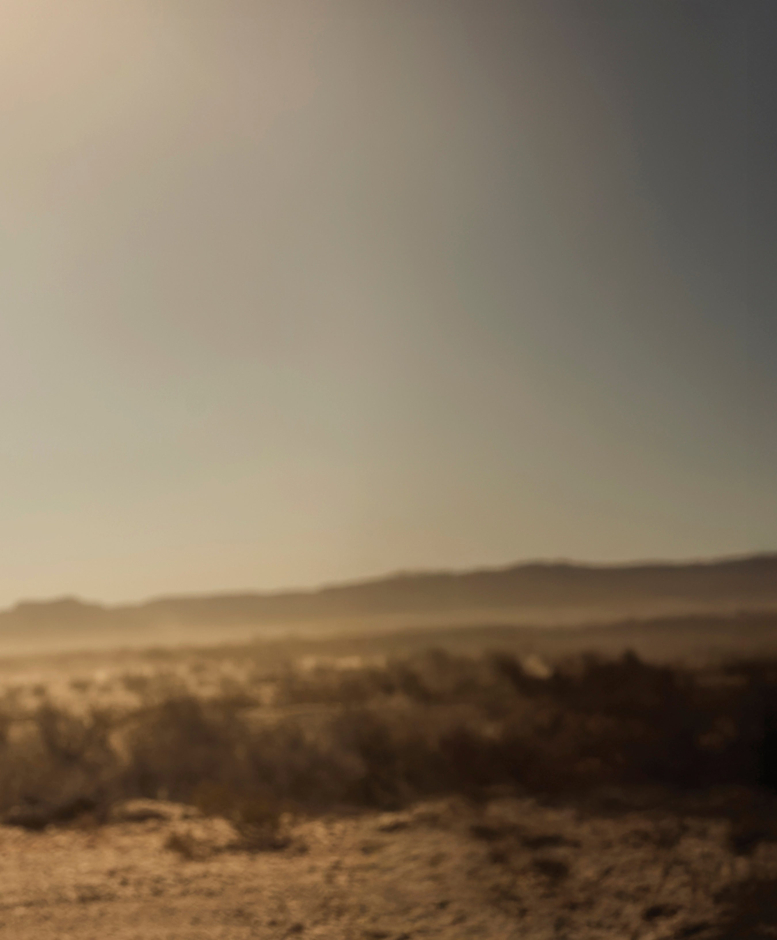 A soft-focus, hazy landscape of a dusty, sunlit desert or prairie with mountains in the background; an intentional transitional background image.