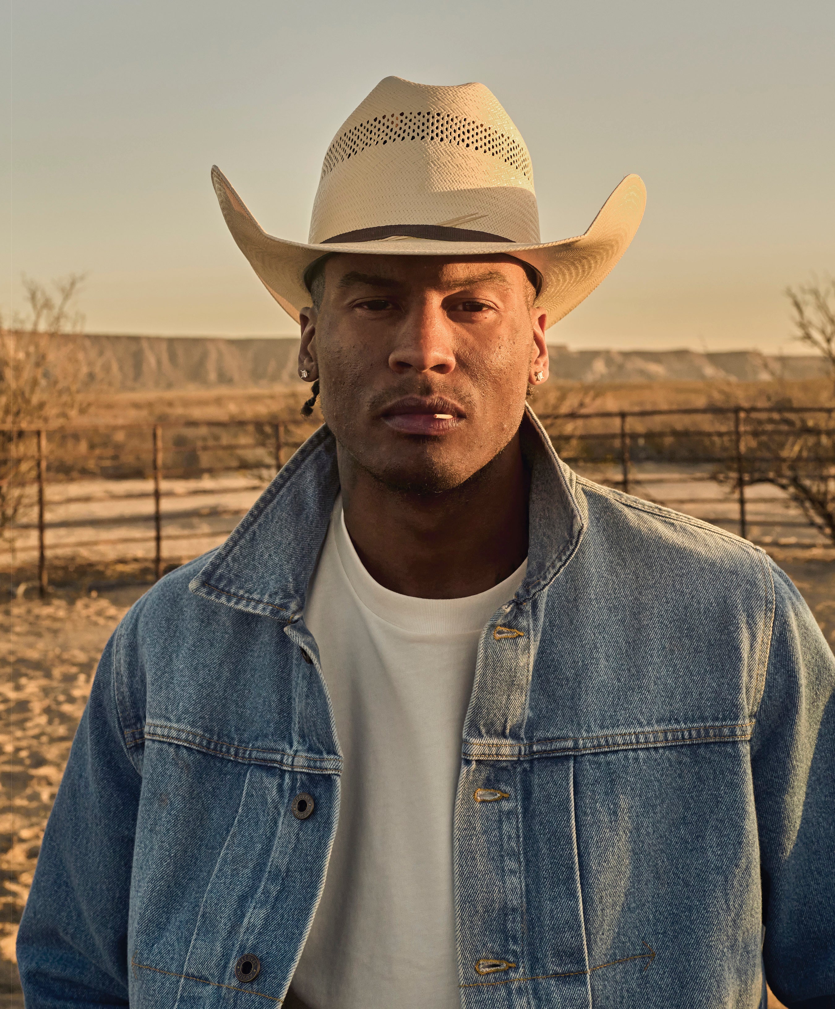 A close-up portrait of a man in a light-colored, perforated straw cowboy hat and a denim jacket over a white t-shirt, standing outdoors in a dry, fenced, desert-like environment at sunset.