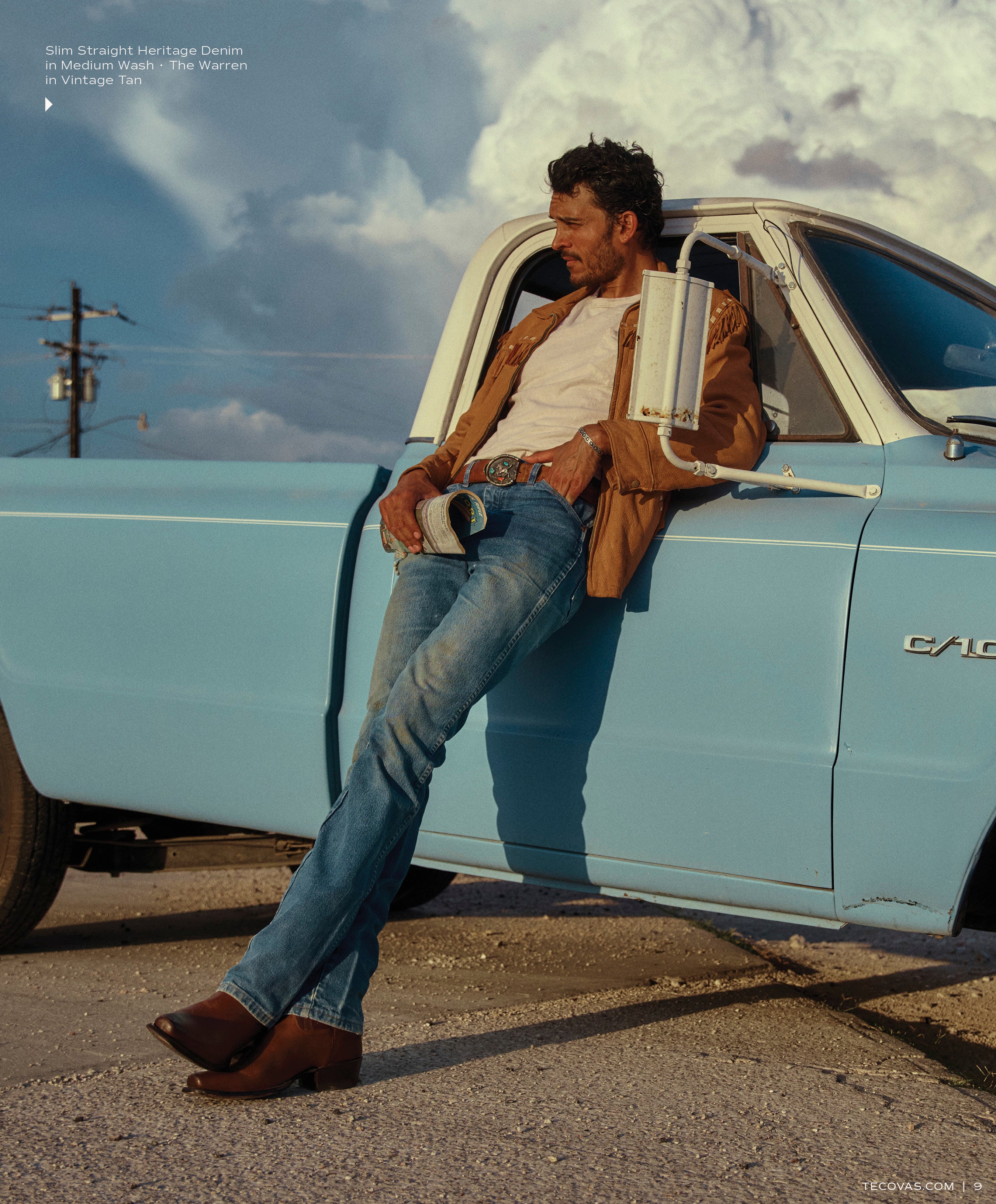 A man with a jacket and cowboy boots is leaning against the fender of a light blue vintage pickup truck, wearing Slim Straight Heritage Denim in Medium Wash - The Warren in Vintage Tan boots.