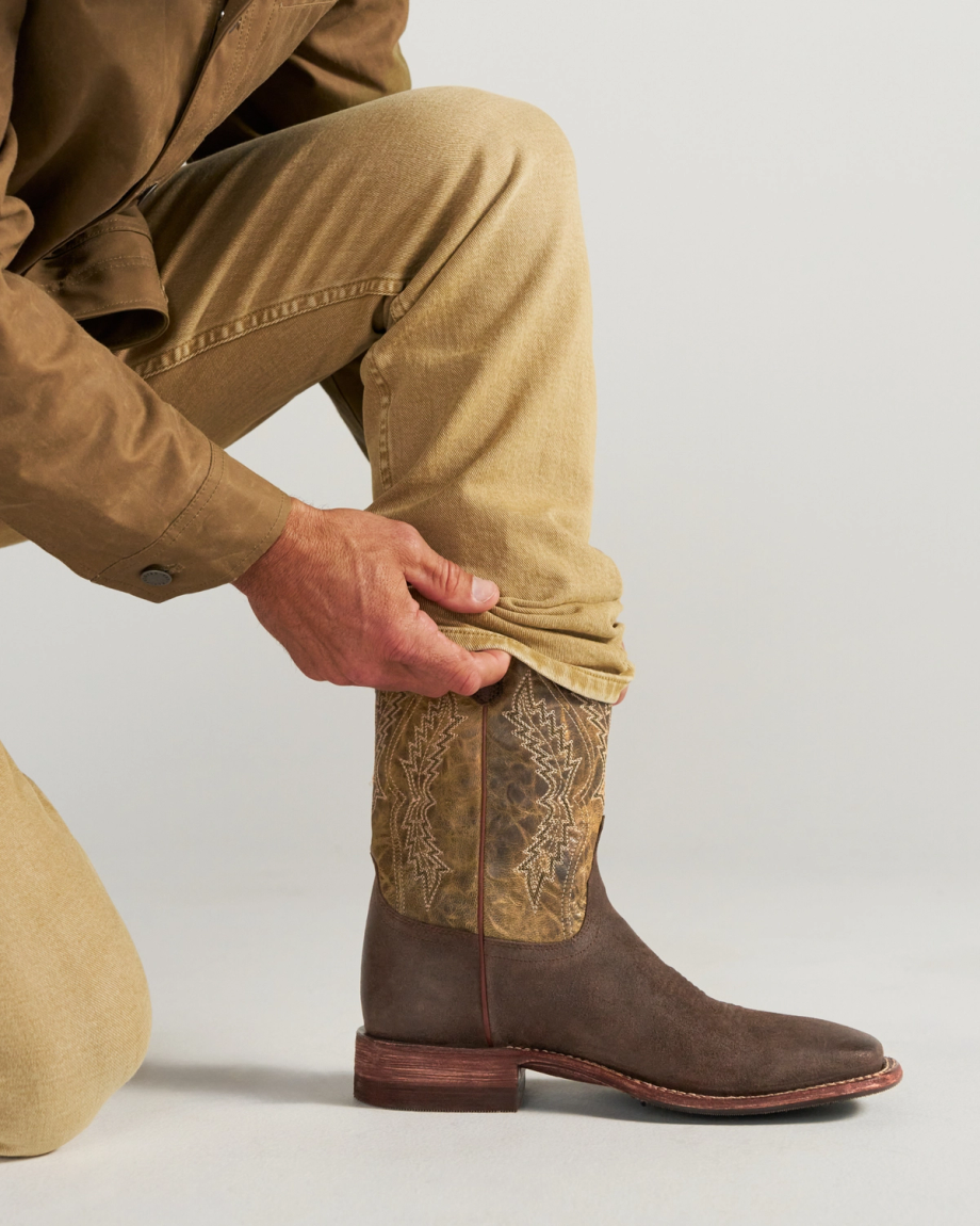 The Doc in Moss Suede. A man wearing tan pants and a brown jacket kneels on a white background, adjusting the hem of his pants over a moss suede cowboy boot with decorative stitching.