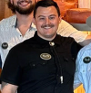 Three people standing together, two partially visible. Central figure wearing a black shirt with a name badge and bolo tie. All are smiling and appear to be in a casual indoor setting.