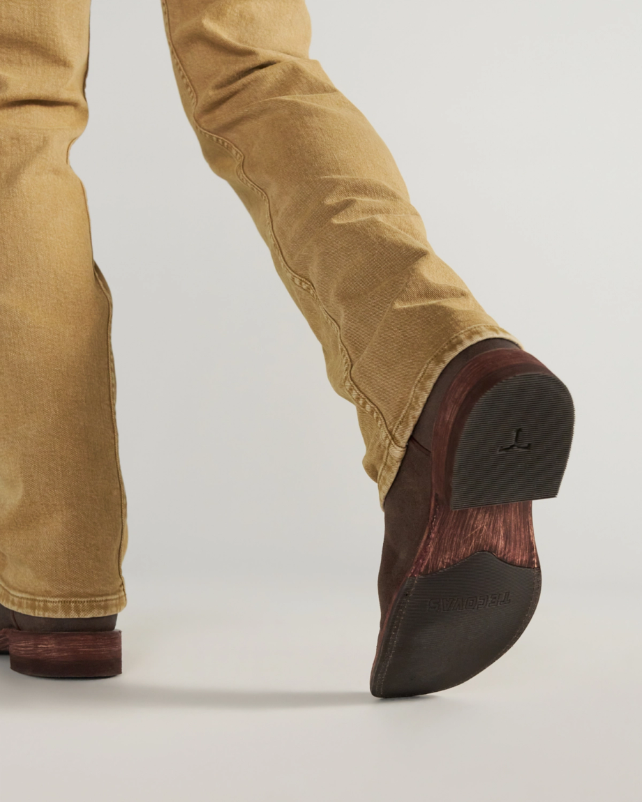 The Doc in Moss Suede. A close-up view of the underside of a Tecovas Moss Suede cowboy boot worn with tan jeans, focusing on the dark brown rubber sole and stacked heel with the brand logo visible.