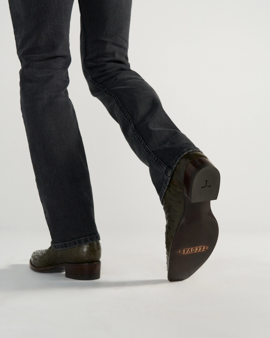 The Jack in Dark Forest Ostrich. Person wearing dark jeans and textured leather boots—crafted from premium Dark Forest Ostrich leather—with one foot lifted to show the sole, which has "TECOYAS" printed on it.
