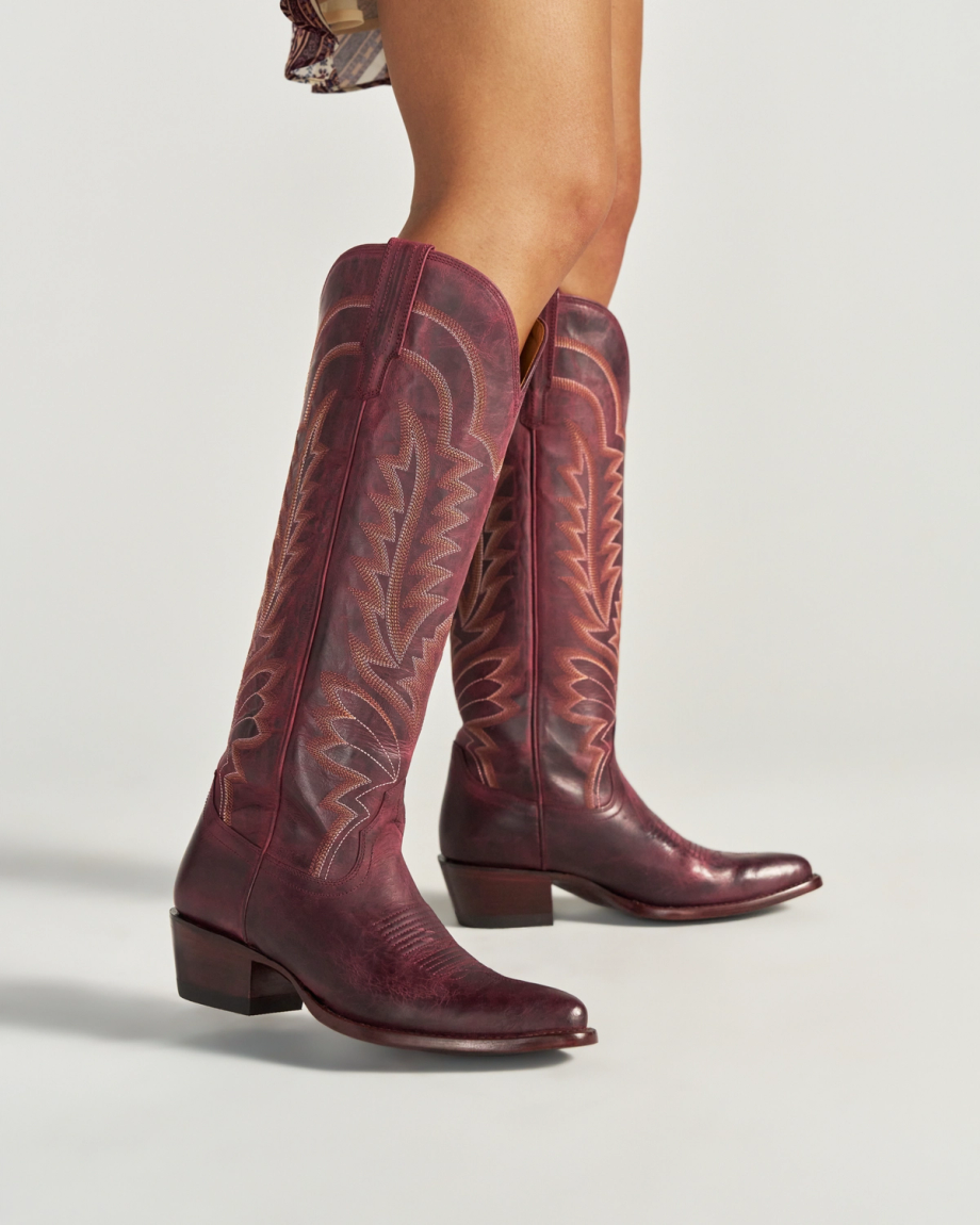 Person wearing tall, dark red cowboy boots with decorative stitching—reminiscent of a bold wine hue—standing on a white background. Only the lower legs are visible, evoking the spirited style of The Abby Goat collection.