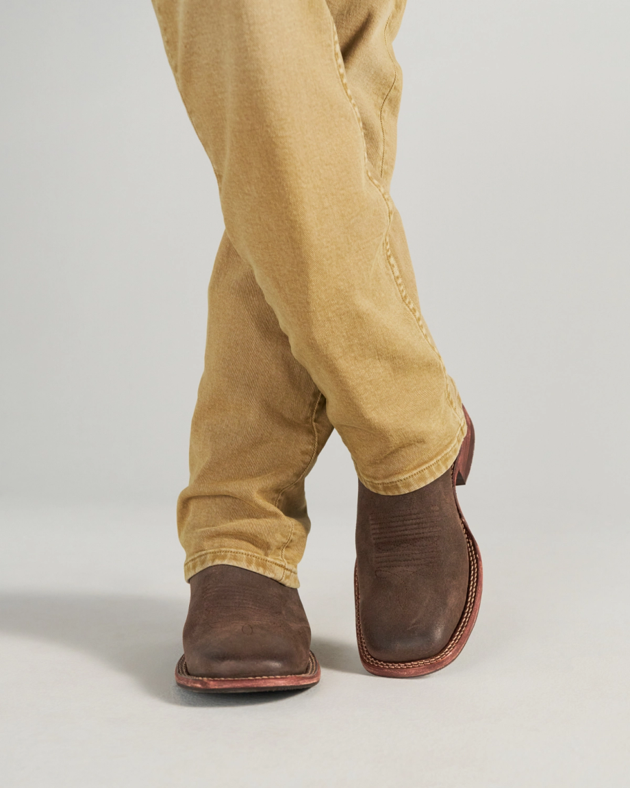 The Doc in Moss Suede. Person wearing light brown pants and Moss Suede cowboy boots, standing with legs crossed on a plain light background.