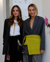 Two women wearing suits with NYFW badges stand side by side indoors; one has long hair and floral pants, the other has a green sash and a flower vase is visible in the background.