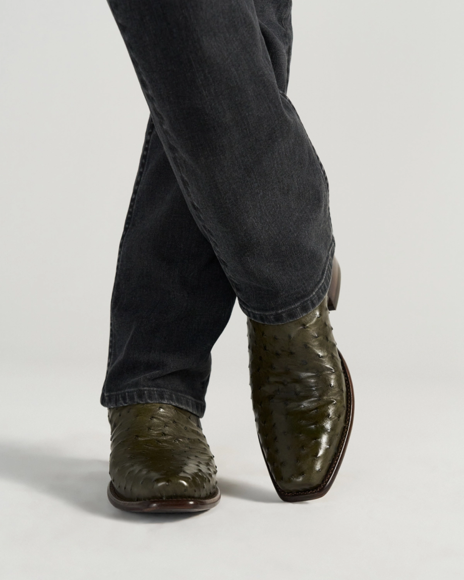 The Jack in Dark Forest Ostrich. Person wearing dark jeans and The Jack Ostrich green ostrich leather cowboy boots, standing with legs crossed against a plain background.