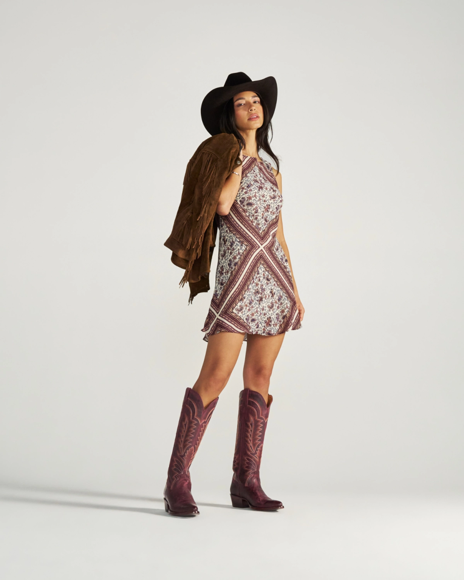 The Abby in Wine Goat. A woman in a cowboy hat, patterned dress, and boots stands holding a brown fringed jacket over her shoulder against a plain background, capturing the free-spirited style of The Abby Goat.