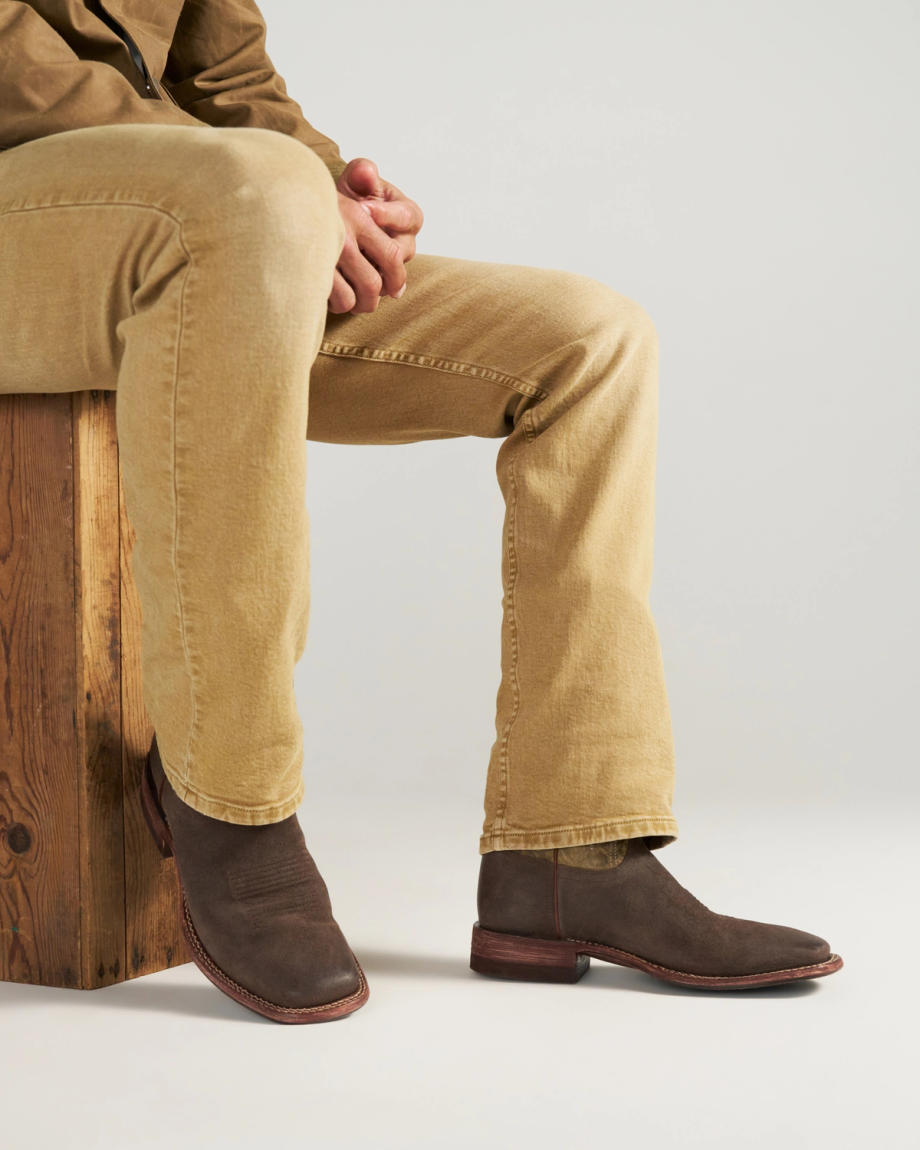 The Doc in Moss Suede. Person sitting on a wooden block, wearing tan pants and brown moss suede boots, with hands resting on knee against a plain light background.