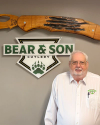 An older man in a white Bear & Son Cutlery shirt stands in front of a Bear & Son Cutlery sign and a large wooden knife display mounted on the wall.