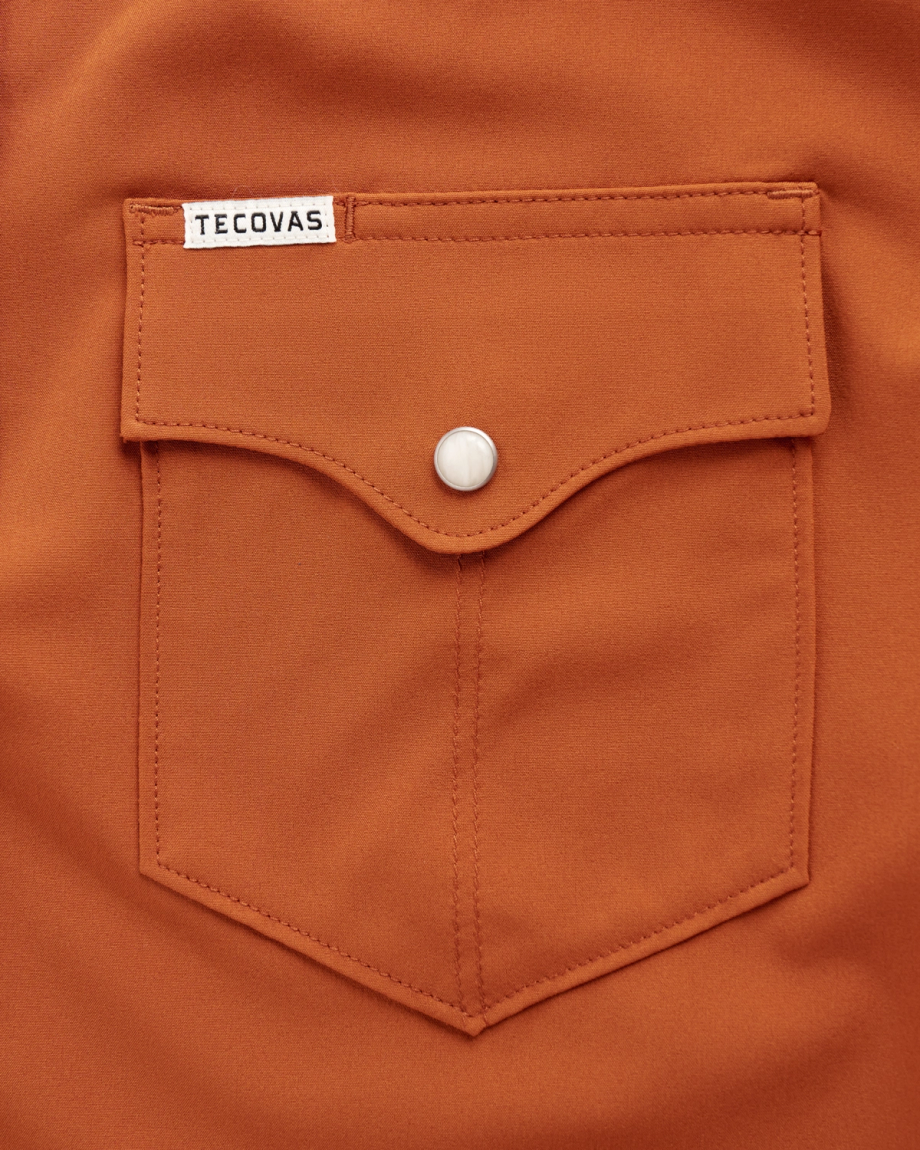 Close-up of a rust-colored shirt pocket with a flap and a snap button. A small white Tecovas label is sewn at the top left of the pocket.
