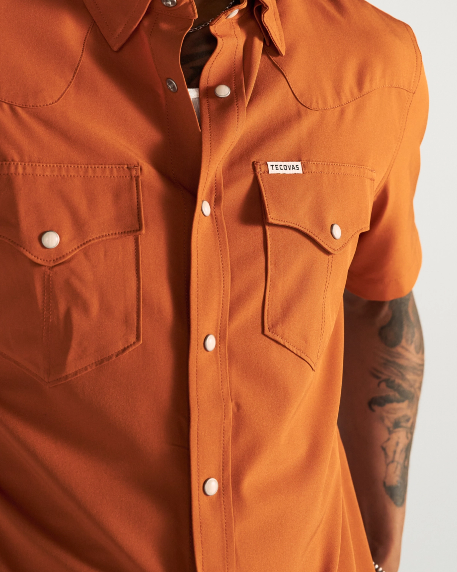 Person wearing an orange short-sleeve button-up shirt with two chest pockets and visible tattoos on their arm.