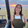 A person smiles in a hot air balloon basket at sunrise, with fields and another balloon visible in the background.