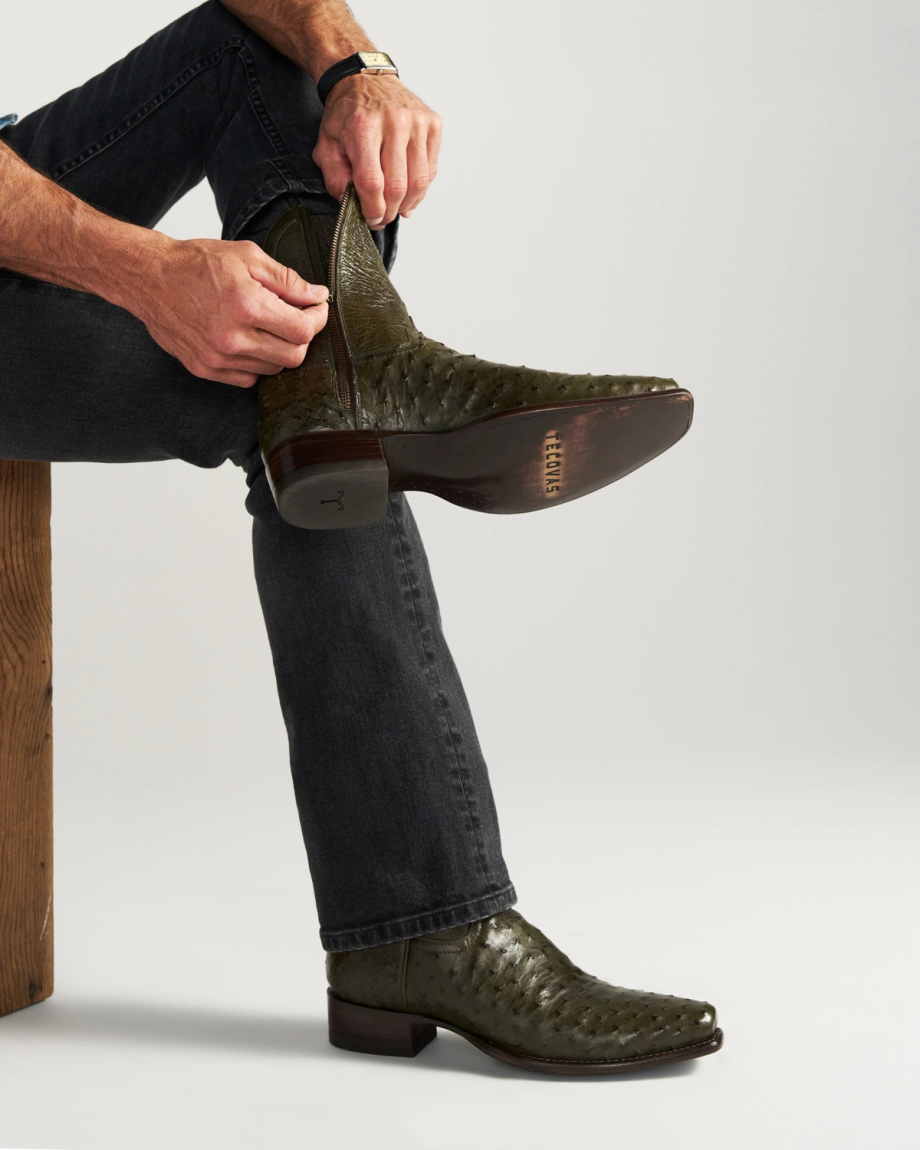 The Jack in Dark Forest Ostrich. A person in dark jeans zips up an olive green textured cowboy boot crafted from luxurious Dark Forest Ostrich leather while sitting on a wooden bench.