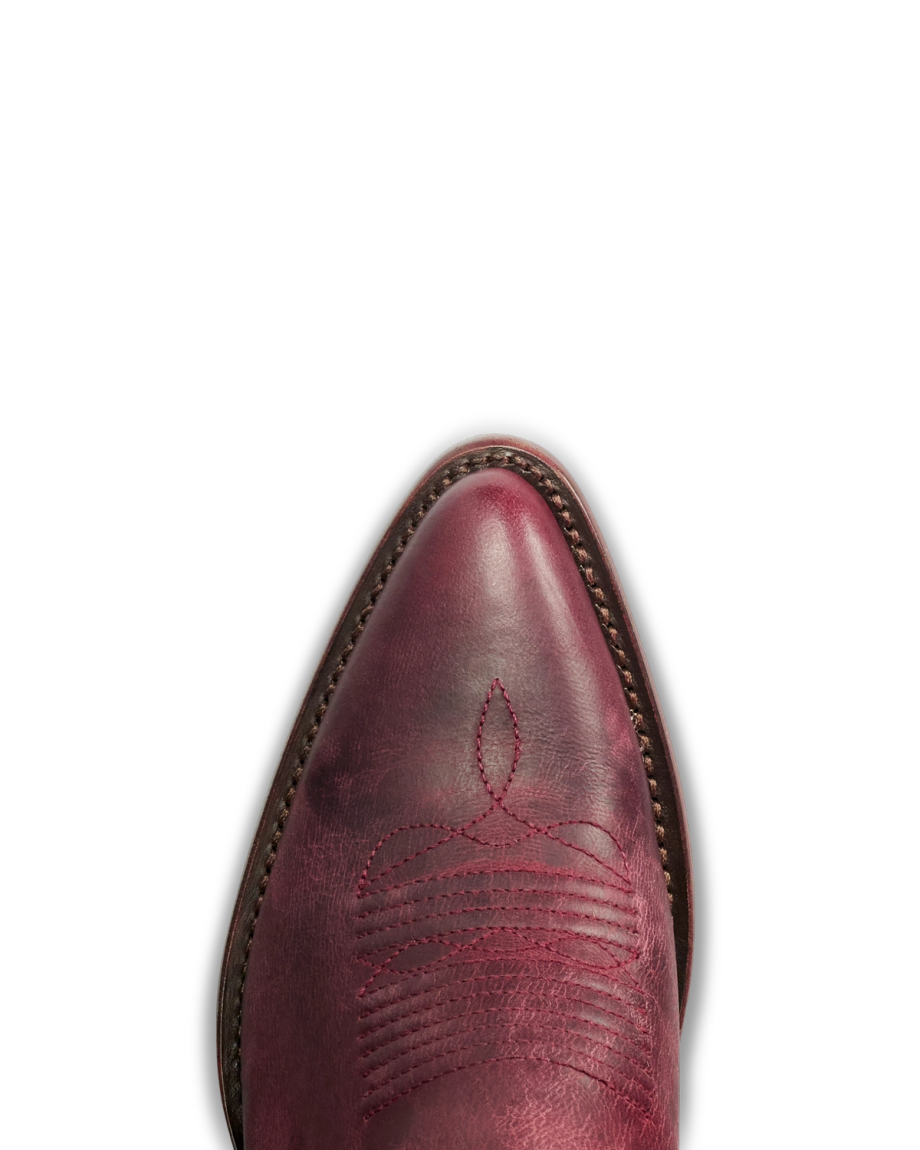 The Abby in Wine Goat. Close-up of the pointed toe of a red leather cowboy boot from The Abby Goat collection, featuring decorative stitching and a brown welt, shown against a white background.