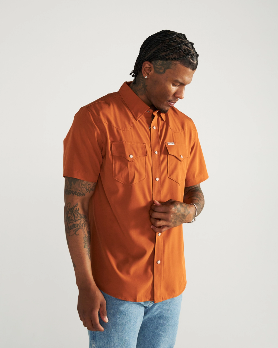 Man wearing an orange short-sleeve button-up shirt with two chest pockets and blue jeans, standing against a plain white background.